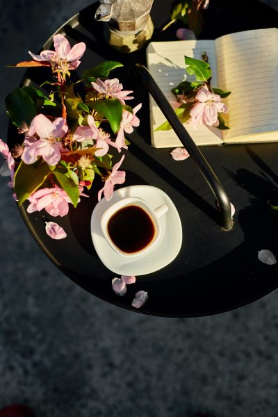 Top view Cup of coffee flowers and notebook on the small black table on terrace at home in a sunny day, outdoor workspace, summer relaxation, coffee break.